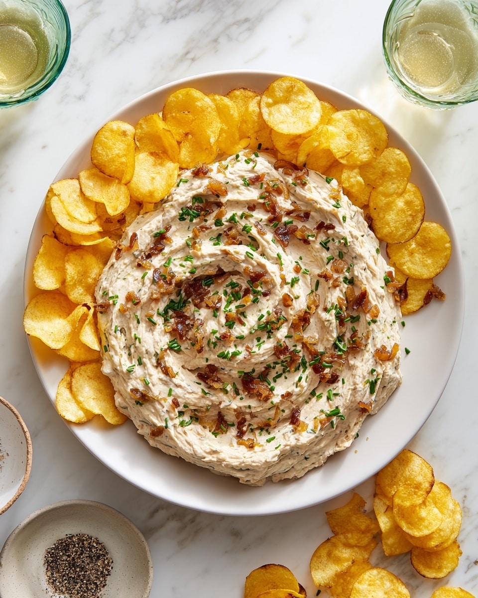 A white plate filled with a creamy dip mixed with browned, cooked onions, giving it a speckled light brown and caramel color texture throughout. The dip is spread roughly but evenly across the plate with chopped green herbs sprinkled on top, adding vibrant green spots. Surrounding the plate are many golden yellow potato chips, some placed under the plate edge and others scattered around. The plate sits on a larger light-colored dish on a white marbled surface. Near the top left corner are two green glasses with ice and a liquid inside, and at the bottom right corner is a small white plate with chopped herbs and a glass container of ground black pepper. photo taken with an iphone --ar 4:5 --v 7