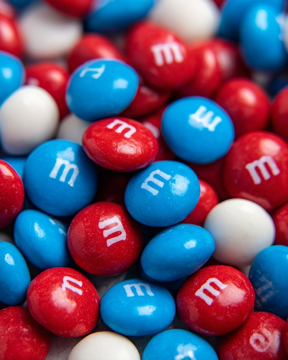 A close-up view of many small round candies, layered tightly together, with three main colors: bright red, white, and blue. Each candy is smooth and shiny with a small white