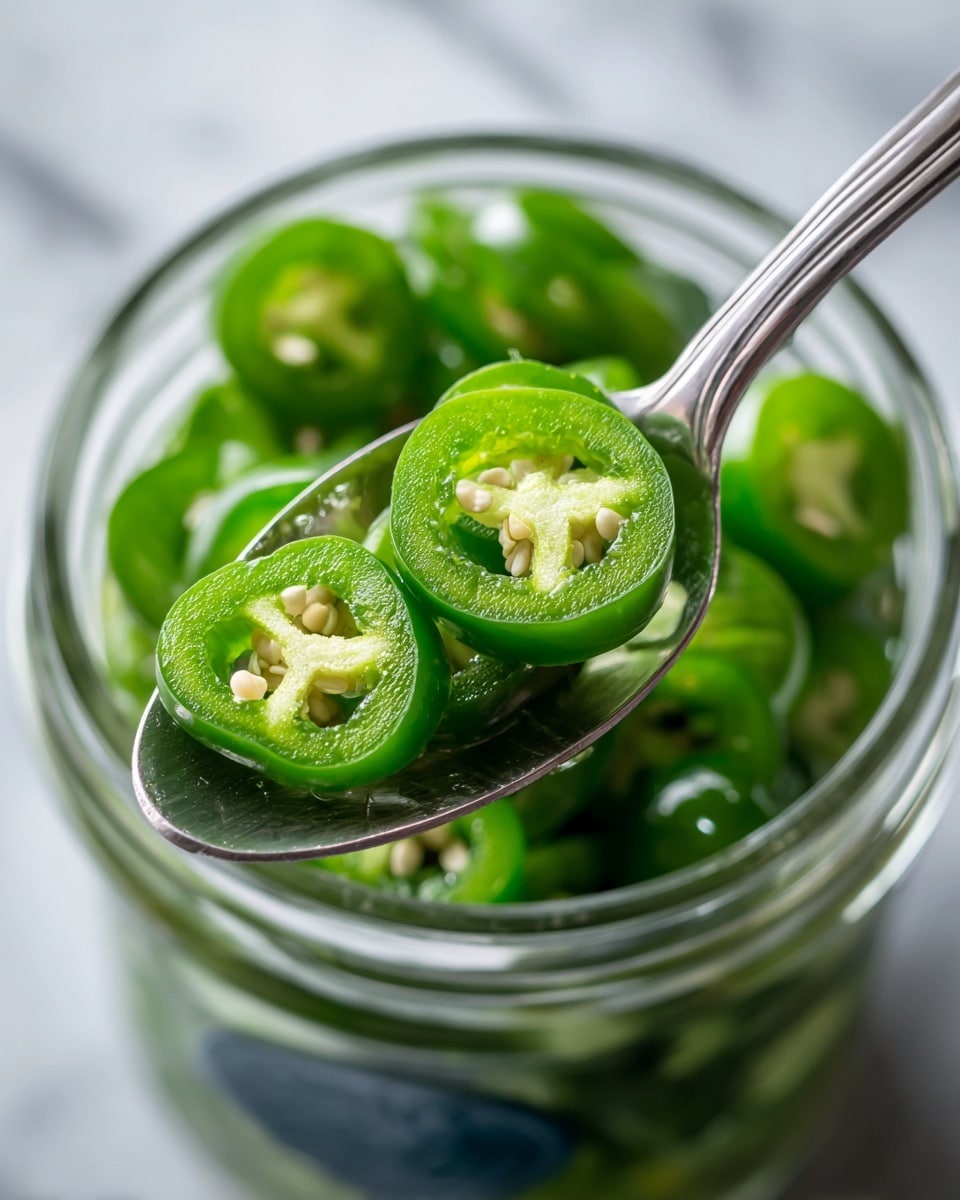 A close-up view of sliced green jalapenos on a shiny silver spoon held above an open glass jar filled with more jalapenos. The jalapeno slices show their pale green color and visible seeds inside, with a slightly wet and glossy texture. The glass jar has a clear rim and a dark blue label faintly visible on the lower part. The background is a white marbled texture, making the green color of the jalapenos stand out. photo taken with an iphone --ar 4:5 --v 7