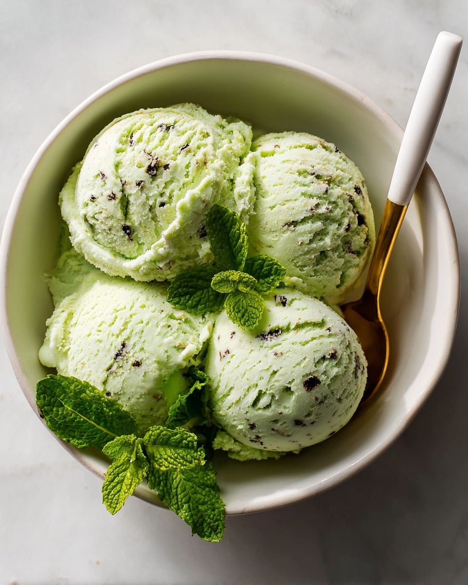 A white bowl holds two large scoops of pale green ice cream speckled with small dark bits, giving it a creamy, textured look. A golden spoon nestles into the right side of the ice cream, partially covered. Fresh bright green mint leaves rest at the bottom left edge of the bowl, adding a pop of color. The bowl sits on a surface with a white marbled texture, bright and clean. photo taken with an iphone --ar 4:5 --v 7