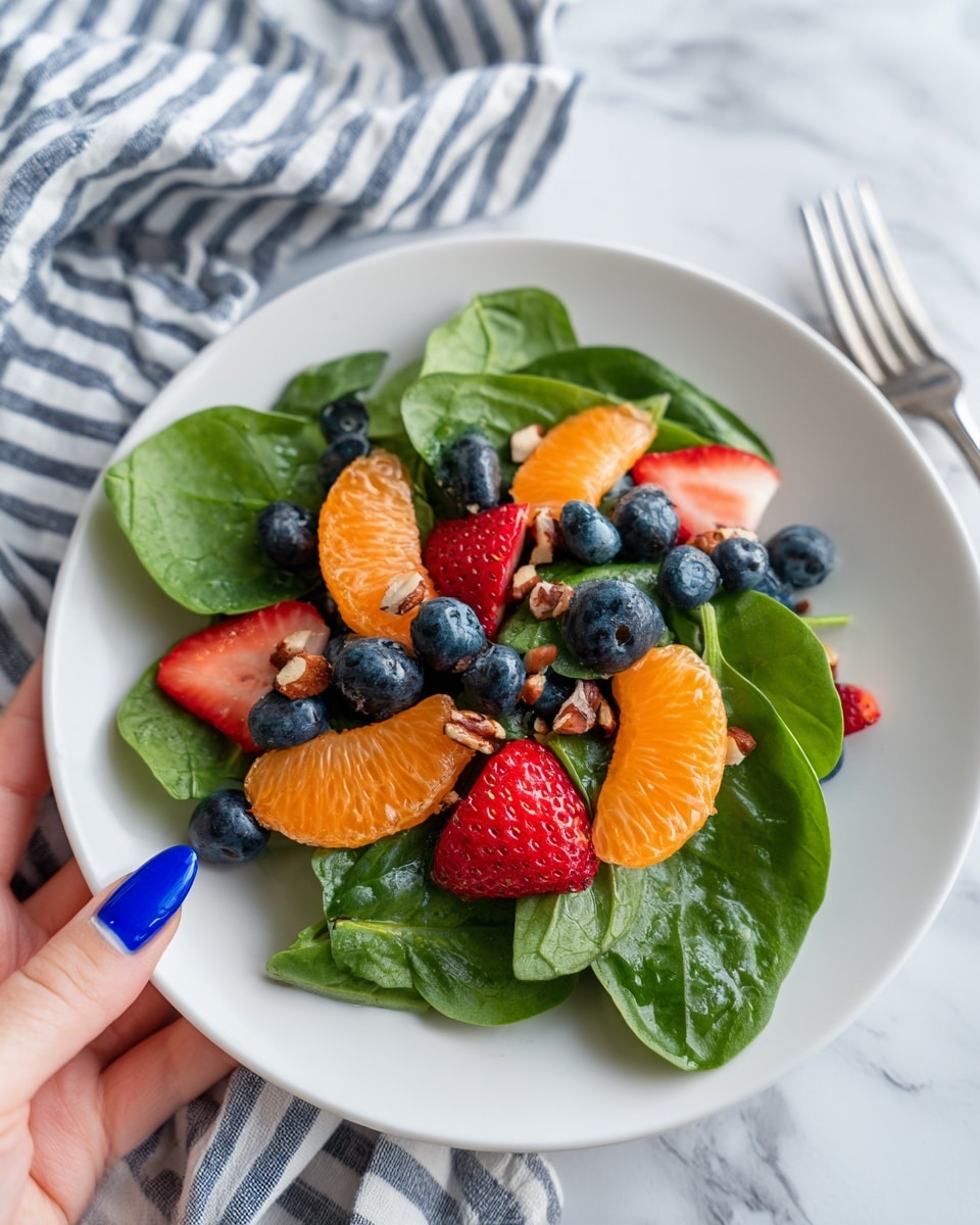 A white bowl filled with a colorful fruit and spinach salad sits on a white marbled surface. The base layer is fresh green spinach leaves with smooth, slightly shiny textures. On top, there are bright orange mandarin slices scattered evenly, their juicy segments distinct and plump. Red strawberry slices are mixed in, showing their seeded surface and soft texture. Blueberries, small and round with a deep purplish-blue color, are sprinkled across the salad. Tiny pieces of chopped nuts add mix of light brown tones and crunchy texture spread over the top. A drizzle of dressing lightly coats the leaves and fruit, adding a shiny wet look. A silver spoon is partly visible on the right side inside the bowl. A woman's hand with light blue nail polish touches the edge of the bowl at the bottom right of the image. A white cloth with thin black stripes folds on the left side near the bowl. Photo taken with an iphone --ar 4:5 --v 7